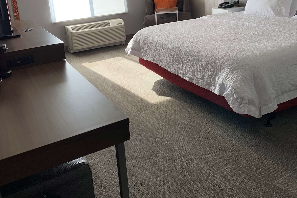 A hotel room with a large bed, a desk, a chair, a window with blinds, a radiator, a lamp, and a painting on the wall.