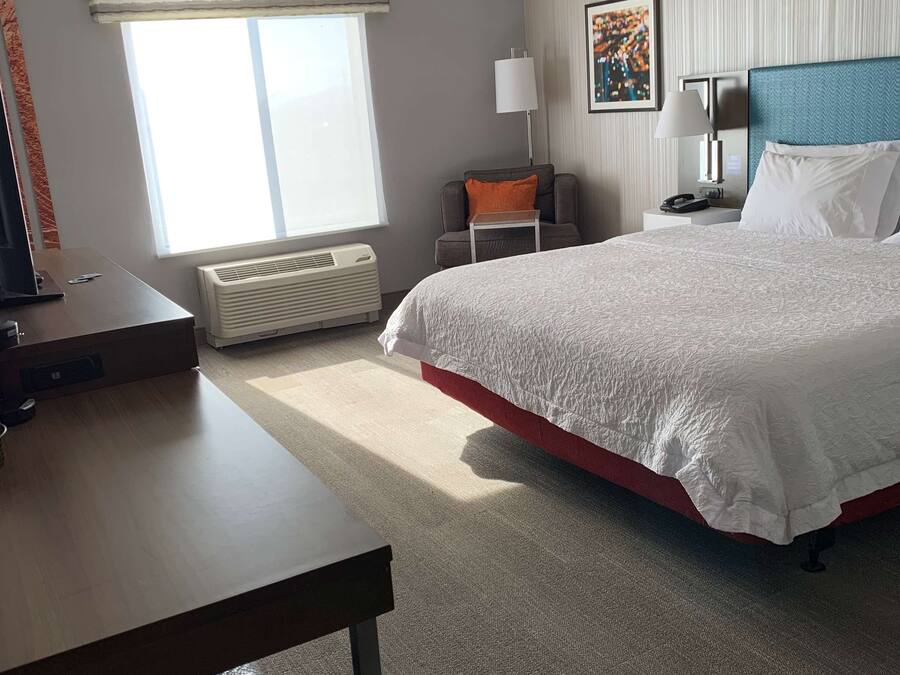 A hotel room with a large bed, a desk, a chair, a window with blinds, a radiator, a lamp, and a painting on the wall.