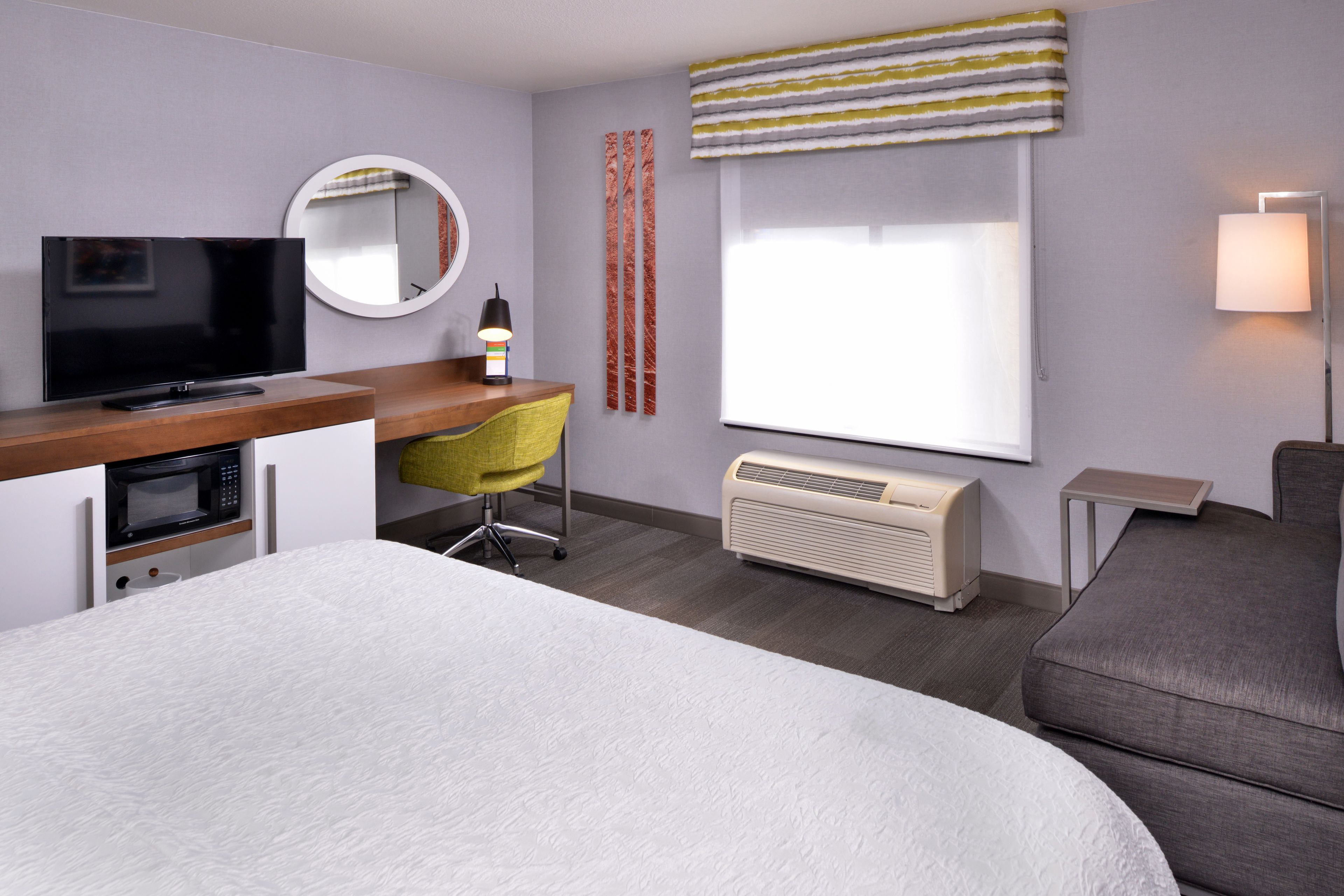 A hotel room with a bed, a desk with a TV, a chair, a mirror, a window with curtains, an air conditioner, and a bedside lamp.