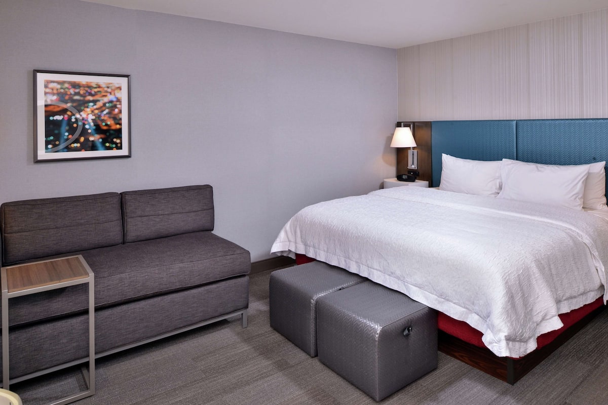 A hotel room with a bed, a grey sofa, a small table, and a wall-mounted lamp.