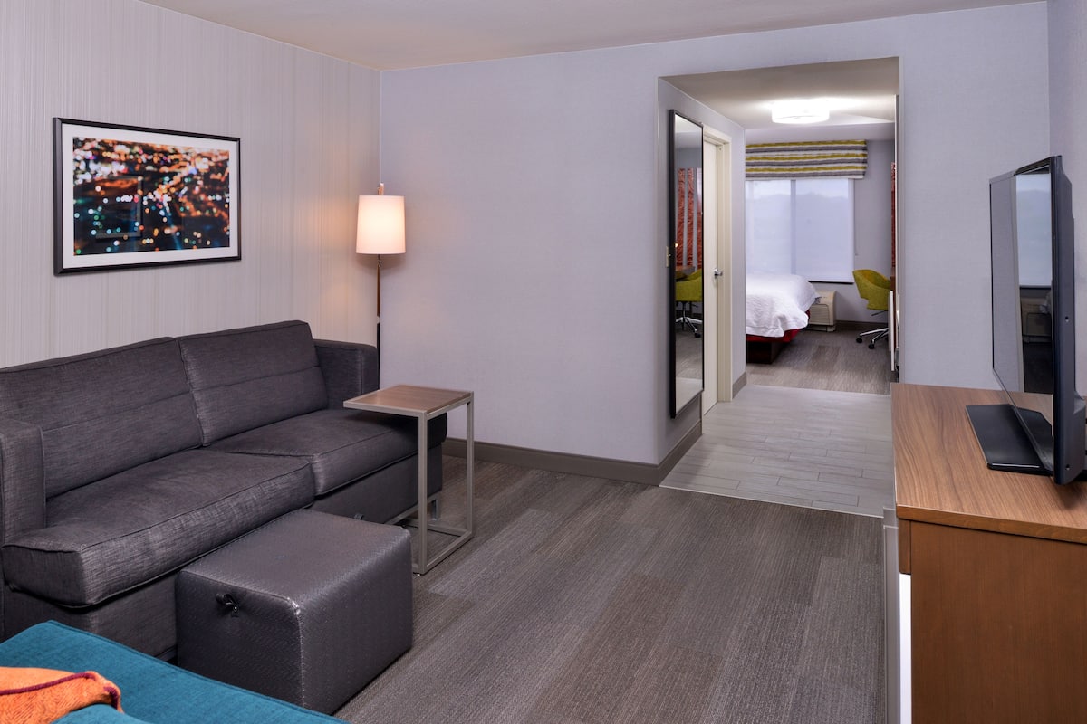 A modern hotel room with a grey sofa, a small table, a TV, and a bed with a yellow chair.