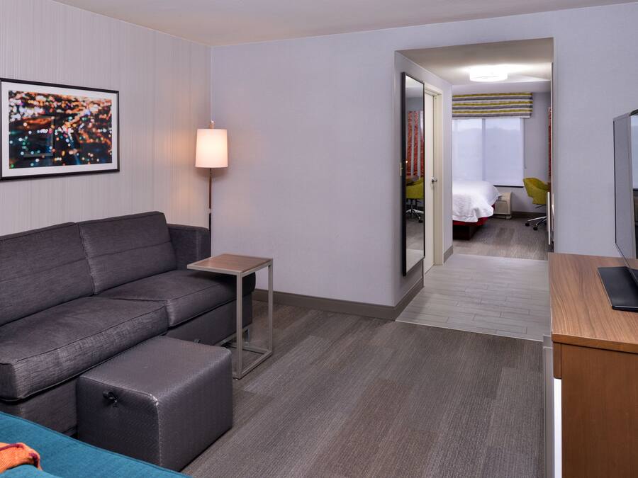 Een moderne hotelkamer met een grijze bank, een kleine tafel, een televisie en een bed met een gele stoel.