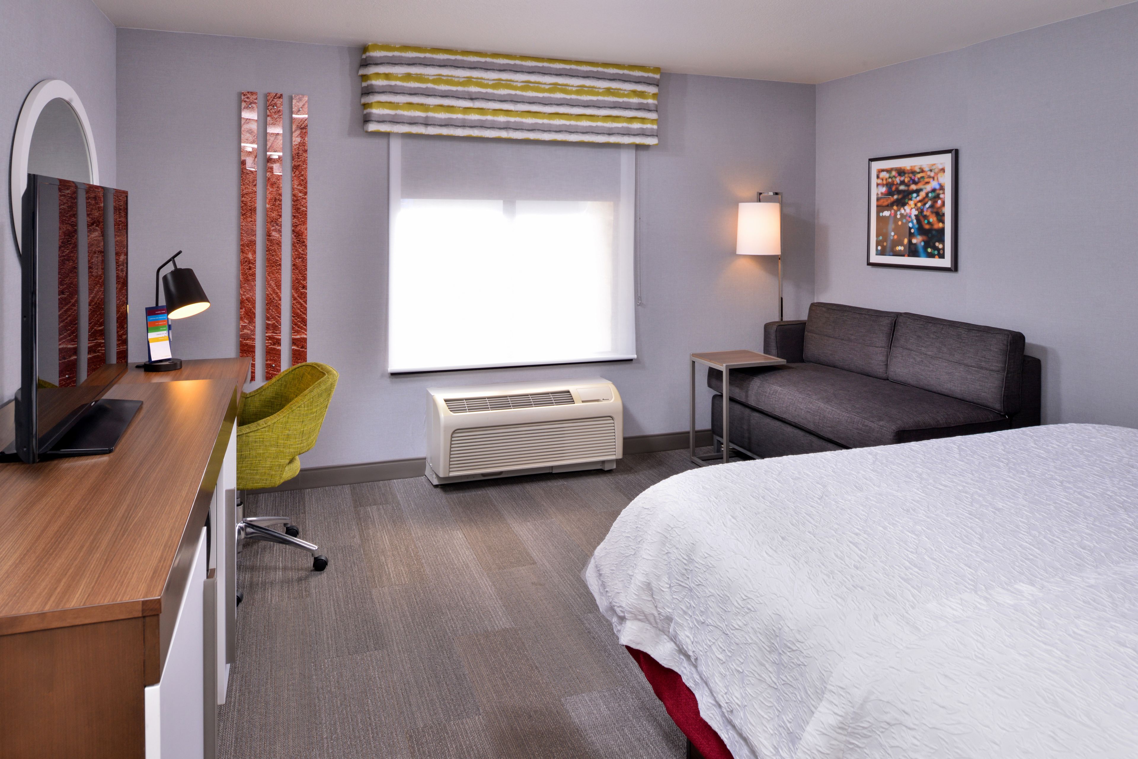 A hotel room with a bed, a desk with a chair, a standing lamp, a heater, and a window with curtains.