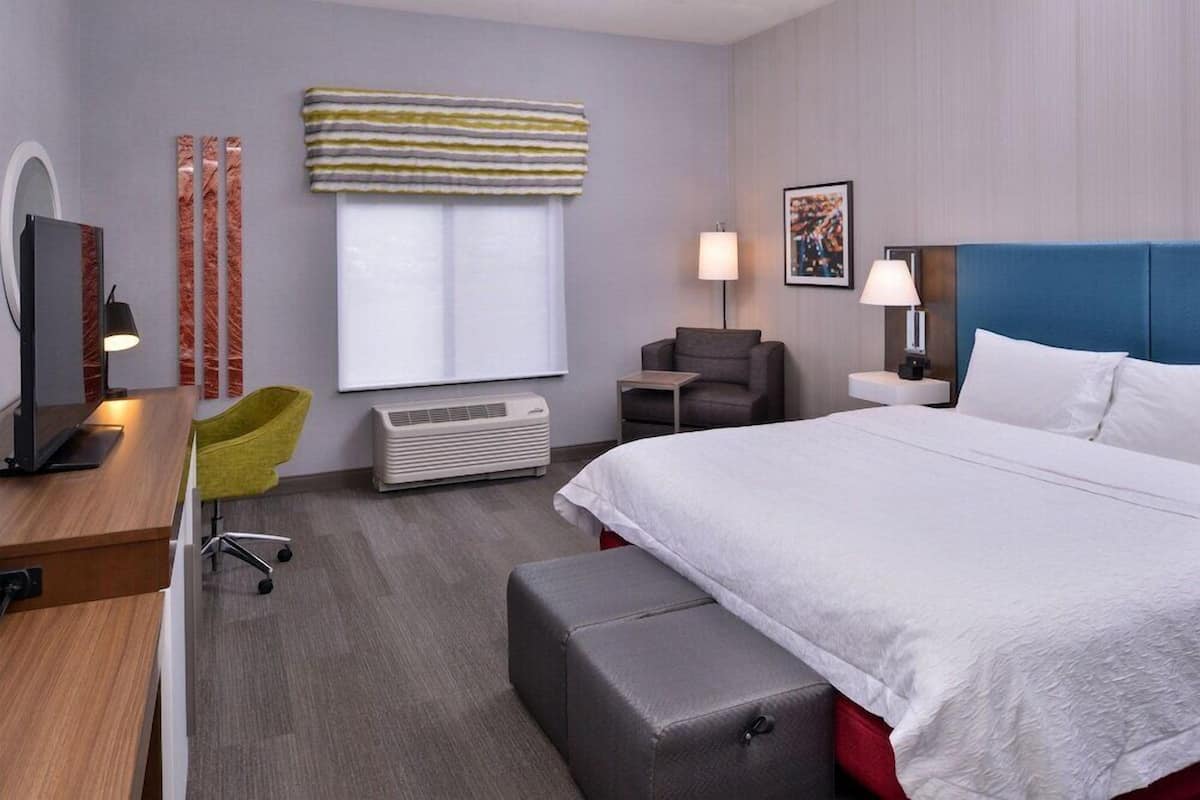 A hotel room with a large bed, a desk with a computer, a chair, a bedside table with a lamp, and a window with curtains.