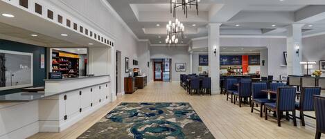 A hotel lobby with a reception desk, seating area, and a view of the kitchen in the background.