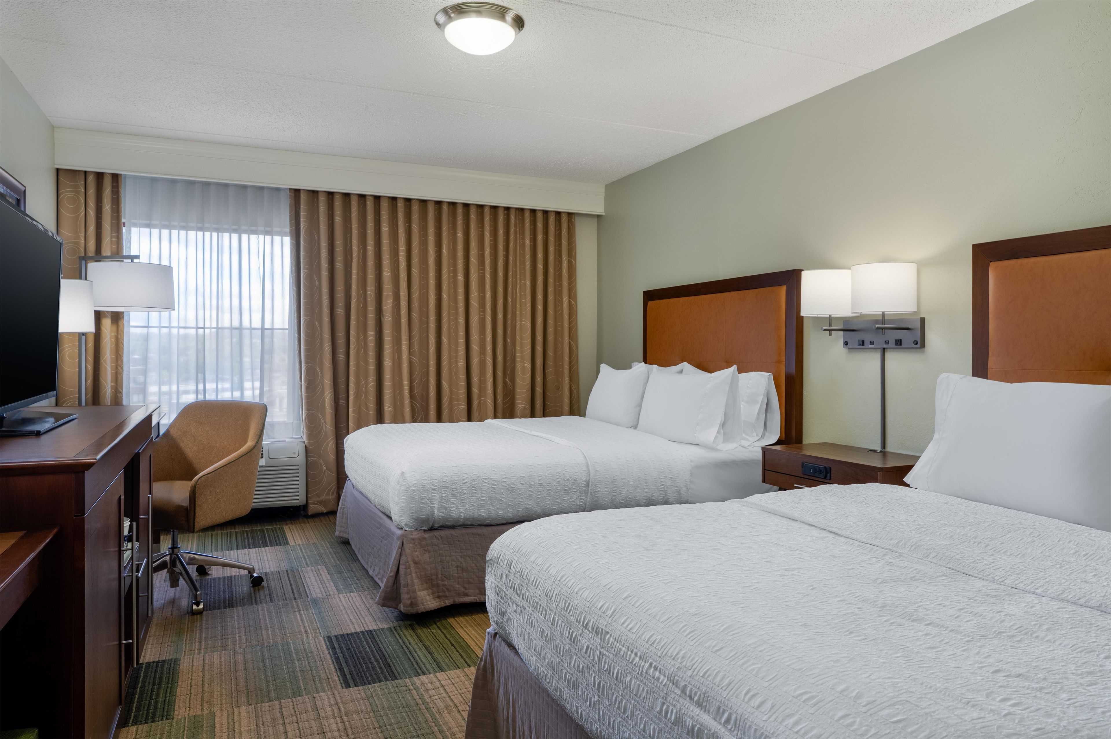 A hotel room with two beds, a desk, a chair, a TV, and a lamp.