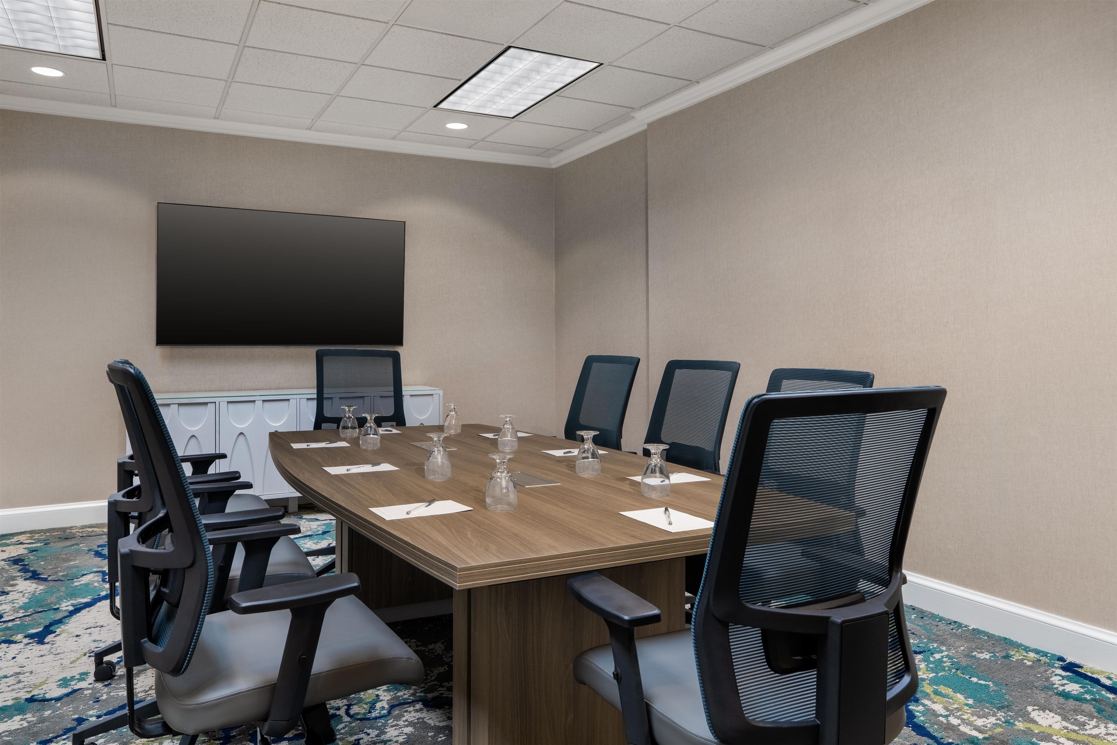 A conference room with a large table, six chairs, a flat-screen TV, and a carpeted floor.
