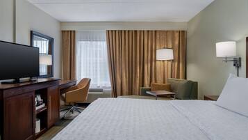 A hotel room with a bed, a desk with a TV, a chair, a small table, and a lamp.