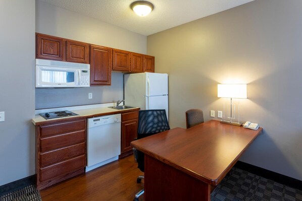 A compact kitchen with wooden cabinets, a microwave, a refrigerator, a dishwasher, and a small dining table with a chair. - GrandStay Hotel & Suites (Ames)