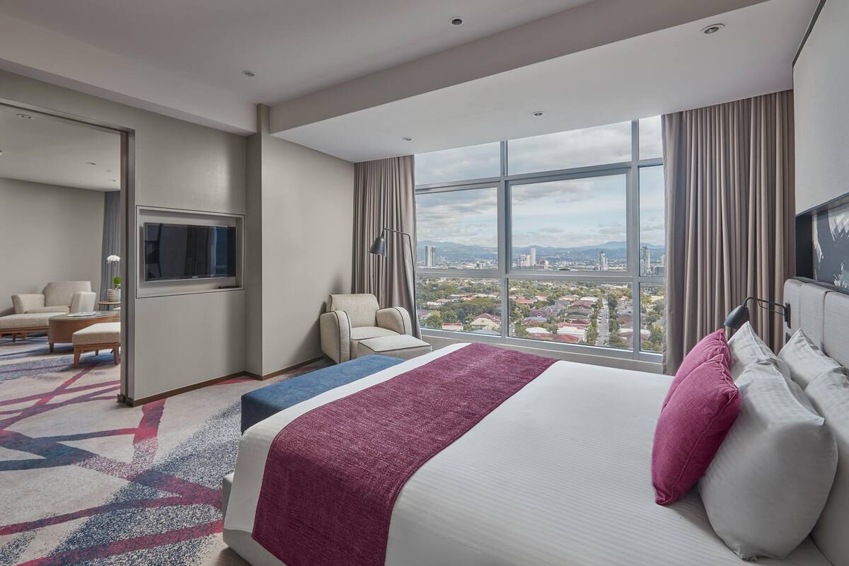 Habitación de hotel con una cama grande, televisión, zona de estar y vista a la ciudad.