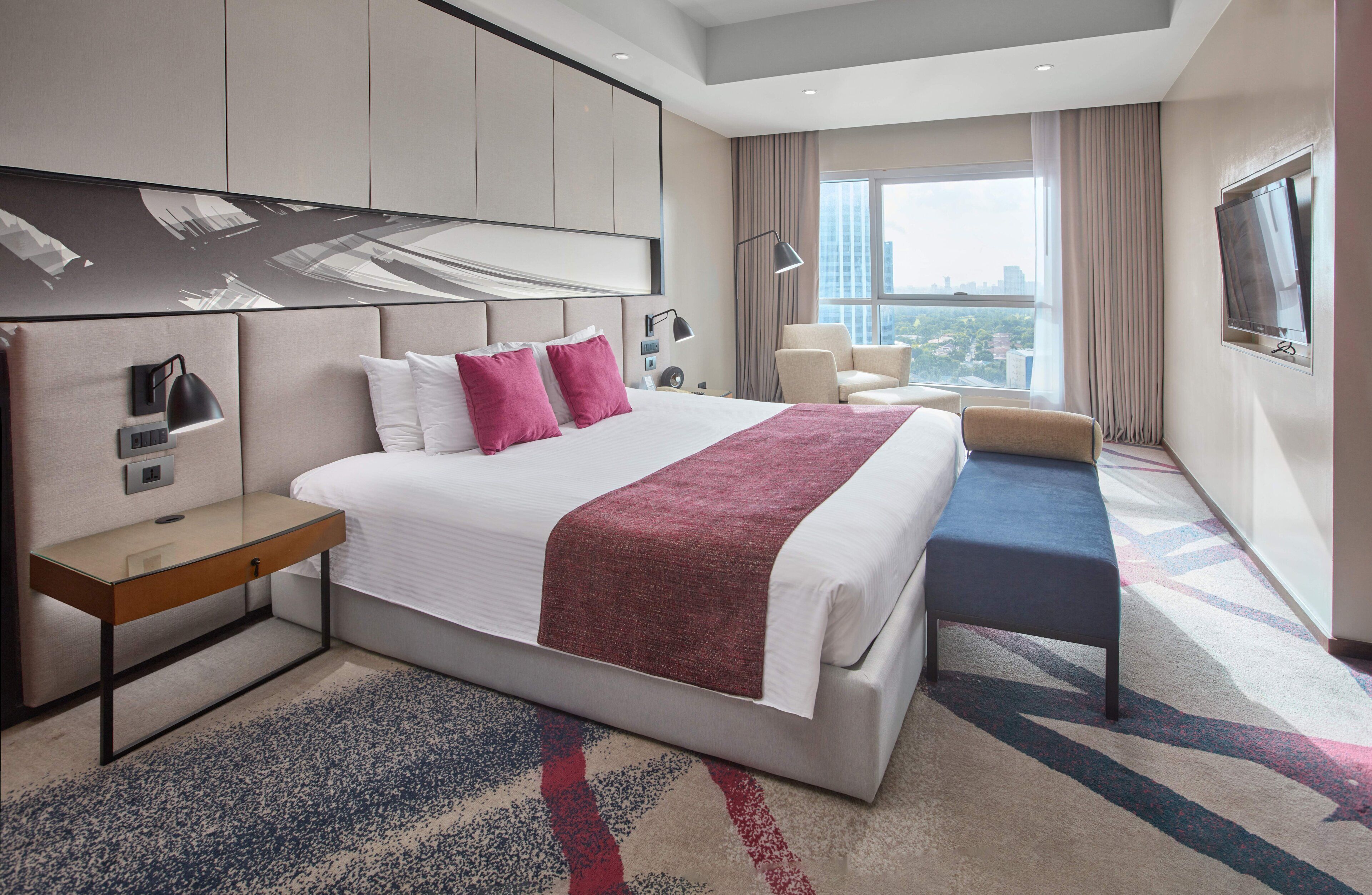 A hotel room with a large bed, bedside tables, a desk, a chair, a TV, and a view of the city.