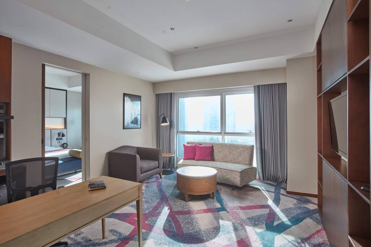 Una habitación de hotel moderna con un ventanal grande, un sofá, una mesita de centro redonda y un escritorio con una silla.
