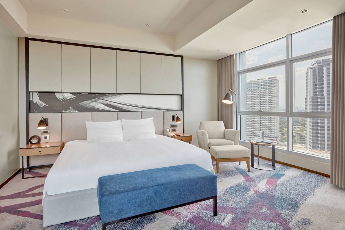 Habitación de hotel con una cama grande, un escritorio, una silla, una mesita y vistas a edificios altos.