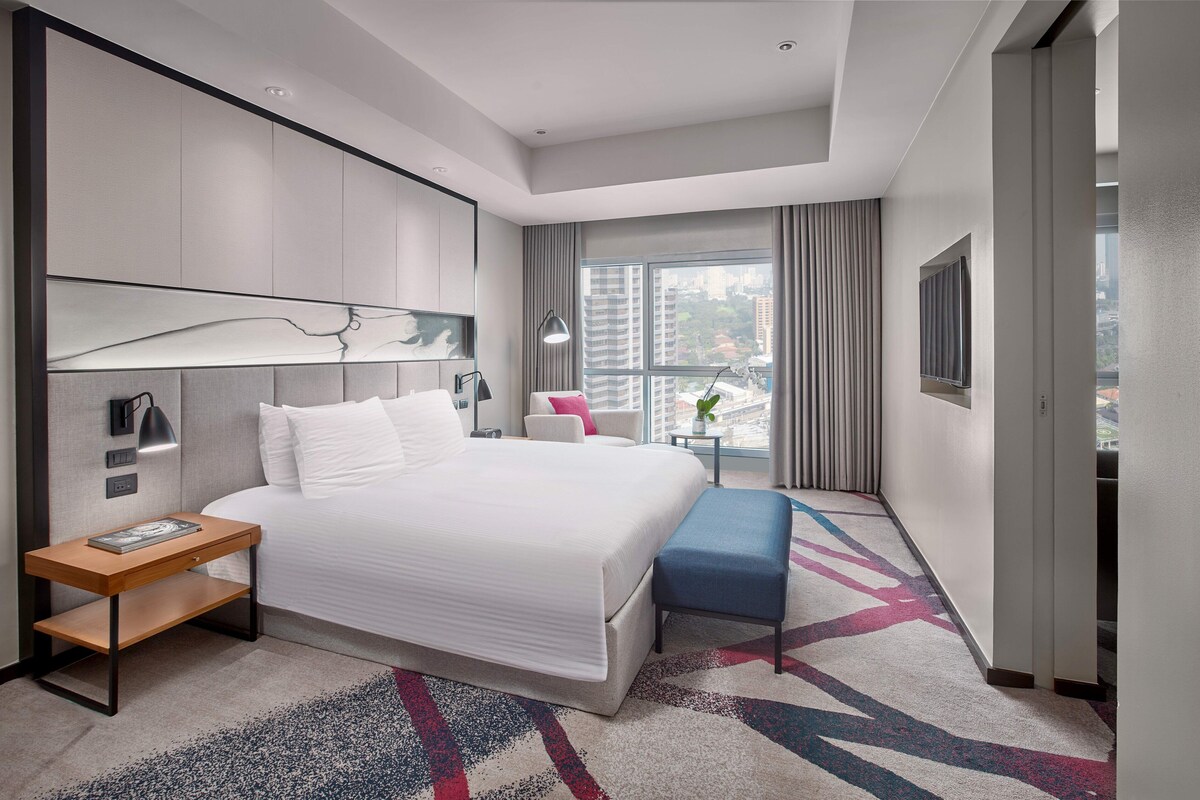 Een moderne hotelkamer met een groot bed, nachtkastje en stadszicht door een groot raam.