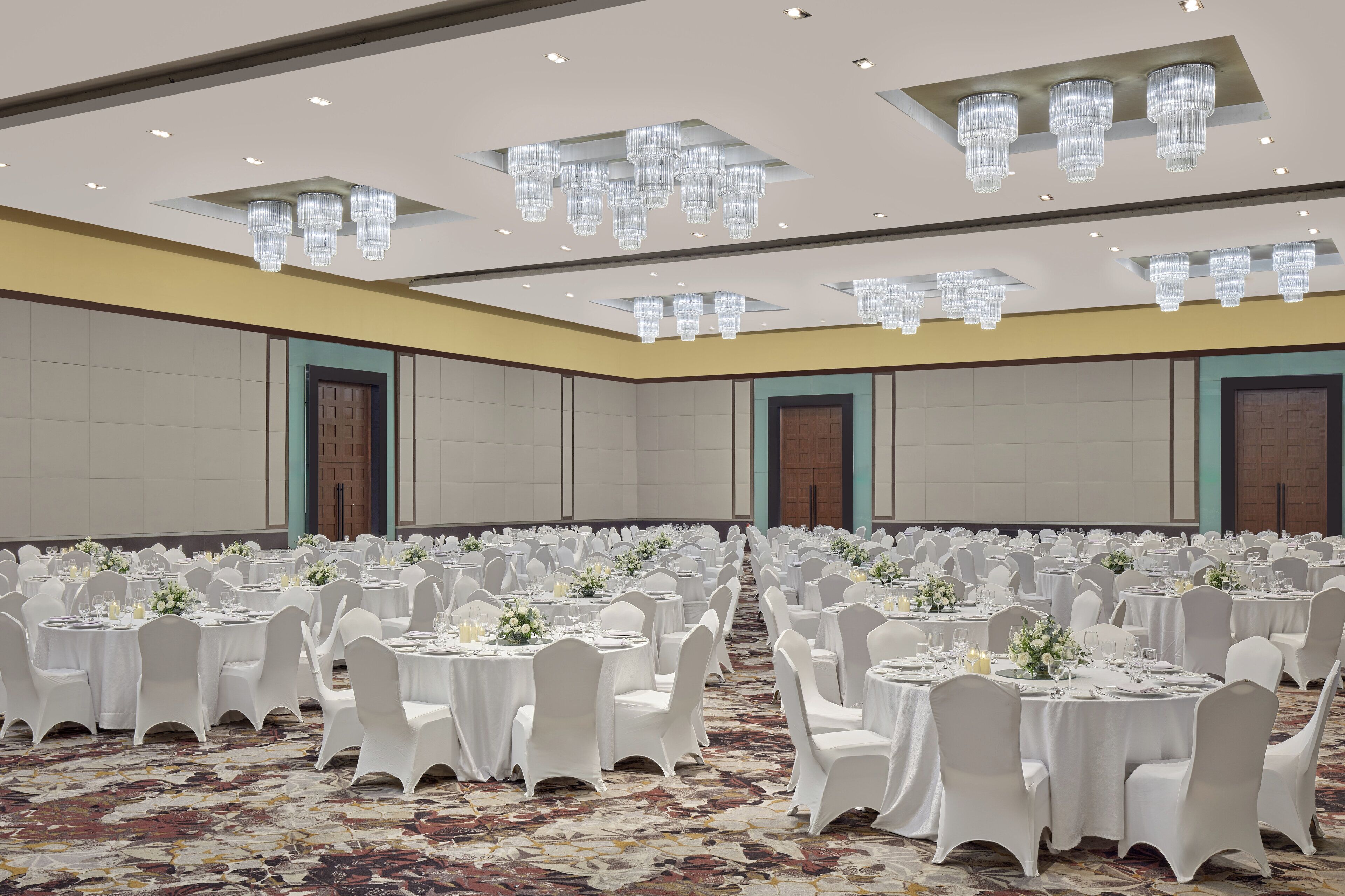 A banquet hall with round tables, white tablecloths, and chairs, featuring a patterned carpet and ceiling chandeliers.