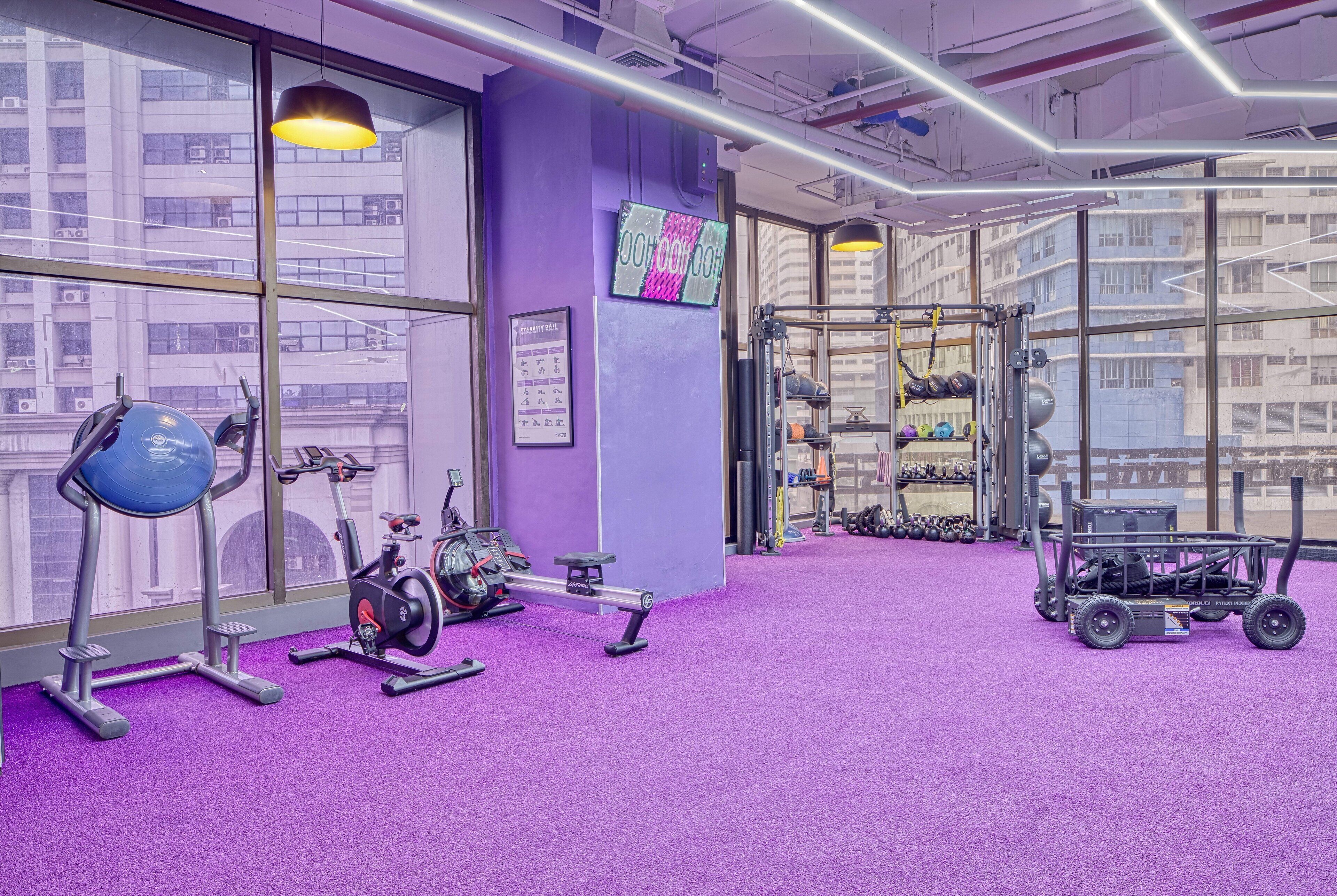 A gym with purple walls, exercise equipment, and a view of buildings through large windows.