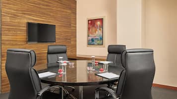 Een vergaderruimte met een ronde tafel, vier zwarte leren stoelen, een flatscreen televisie aan de muur en een kleurrijk schilderij erboven.