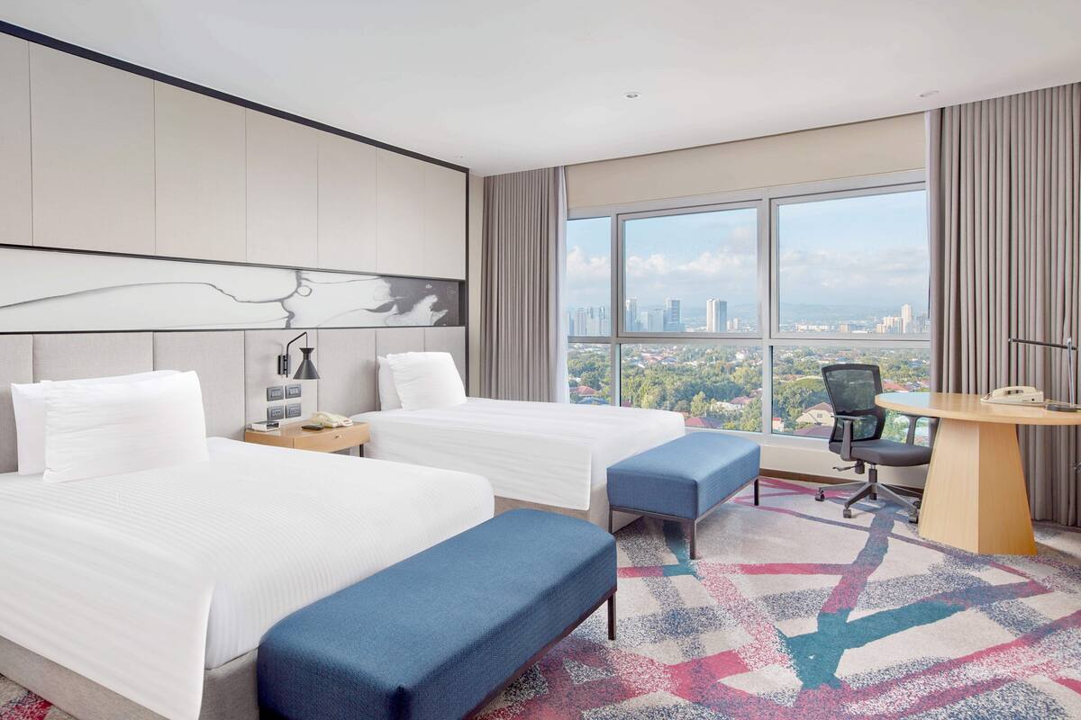 Habitación de hotel con dos camas, un escritorio, una silla y un ventanal con vistas a la ciudad.