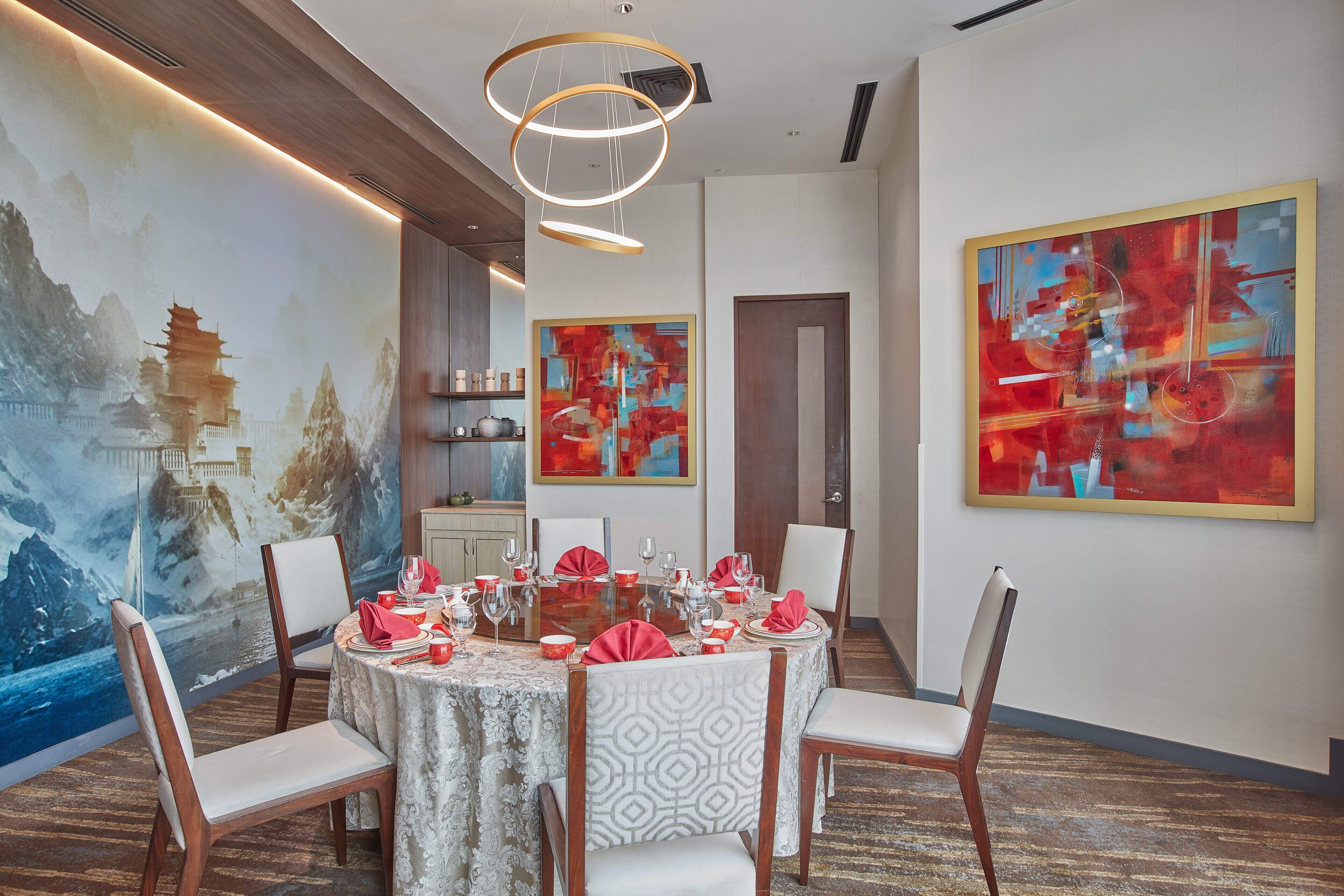 Un coin repas avec une table ronde dressée pour six personnes, entourée de chaises aux motifs d’habillage variés. Un grand tableau abstrait orne le mur à droite, et une fresque représentant une scène asiatique traditionnelle se trouve à gauche.
