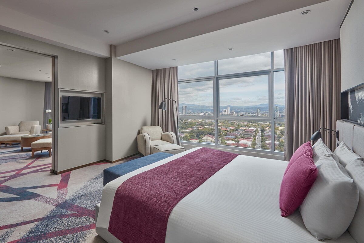 Een hotelkamer met een groot bed, een televisie, een zithoek en uitzicht op een stadsbeeld.