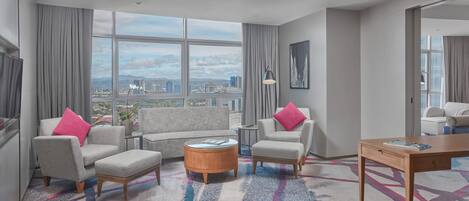 Ein modernes Hotelzimmer mit großem Fenster, grauen Sofas mit rosafarbenen Kissen, einem runden Holztisch und einem Holzbüro.