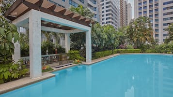 Una piscina con zona cubierta, rodeada de vegetación y edificios altos al fondo.