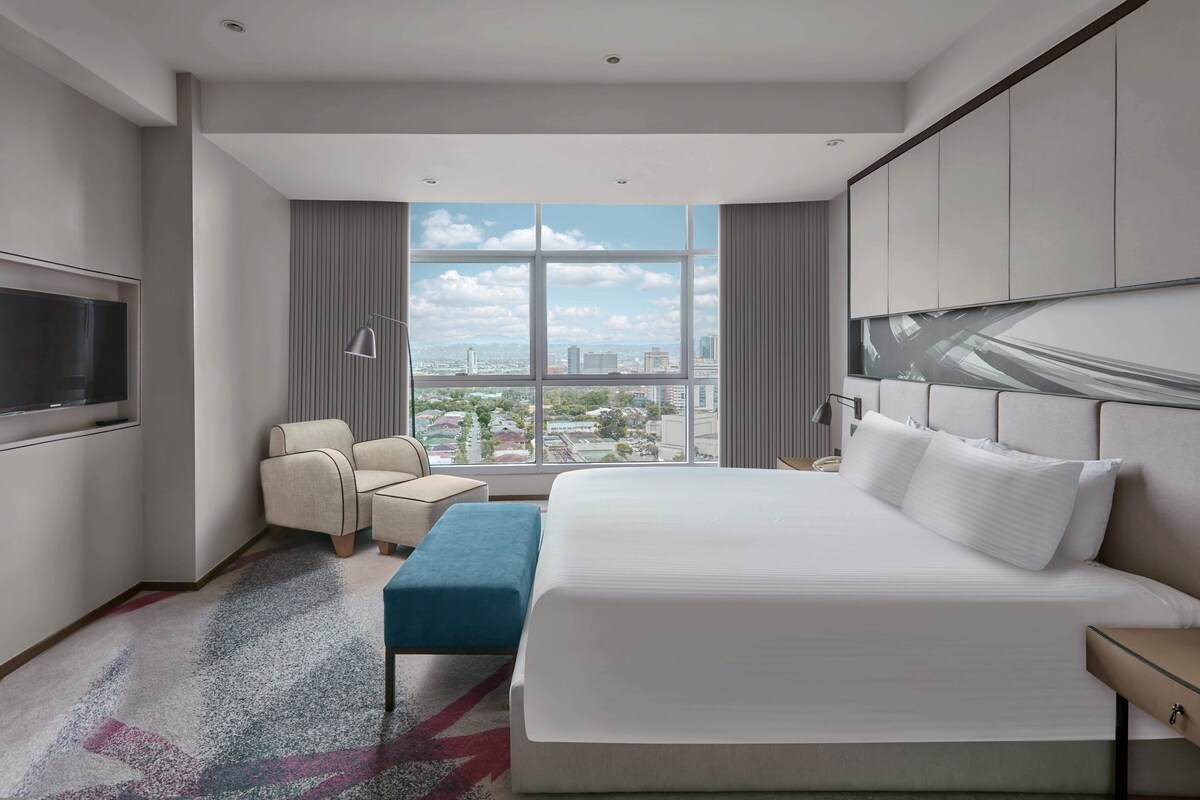 Een moderne hotelkamer met een groot bed, een zithoek met een fauteuil en poef, een flatscreen televisie aan de muur en uitzicht op de stad door grote ramen.