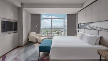 A modern hotel room with a large bed, a seating area with a chair and ottoman, a flat-screen TV mounted on the wall, and a city view through large windows.