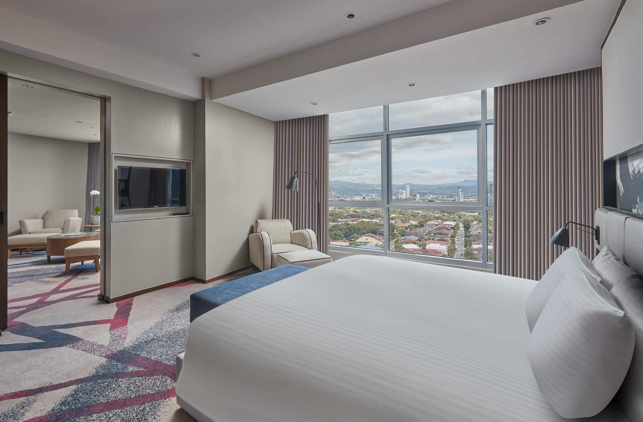Habitación de hotel con una cama grande, televisión, zona de estar con sofá y sillones, y vistas a la ciudad a través de amplios ventanales.