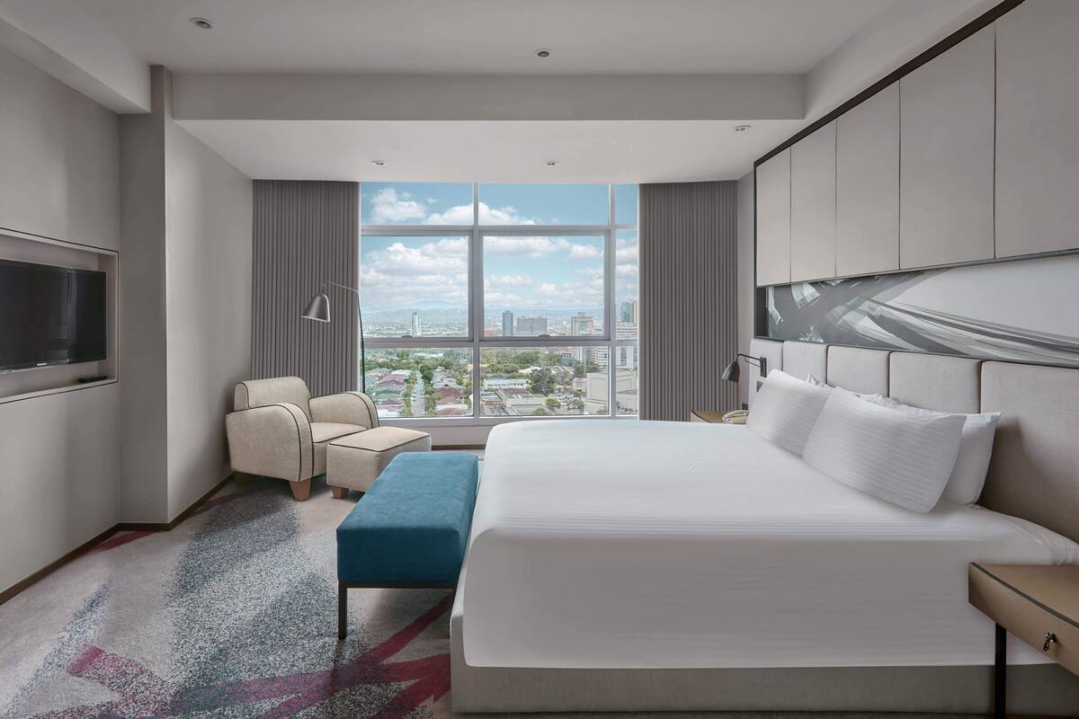 Una habitación de hotel moderna con una cama grande, un área de descanso con una silla y un reposapiés, un televisor de pantalla plana montado en la pared y vistas a la ciudad a través de amplias ventanas.