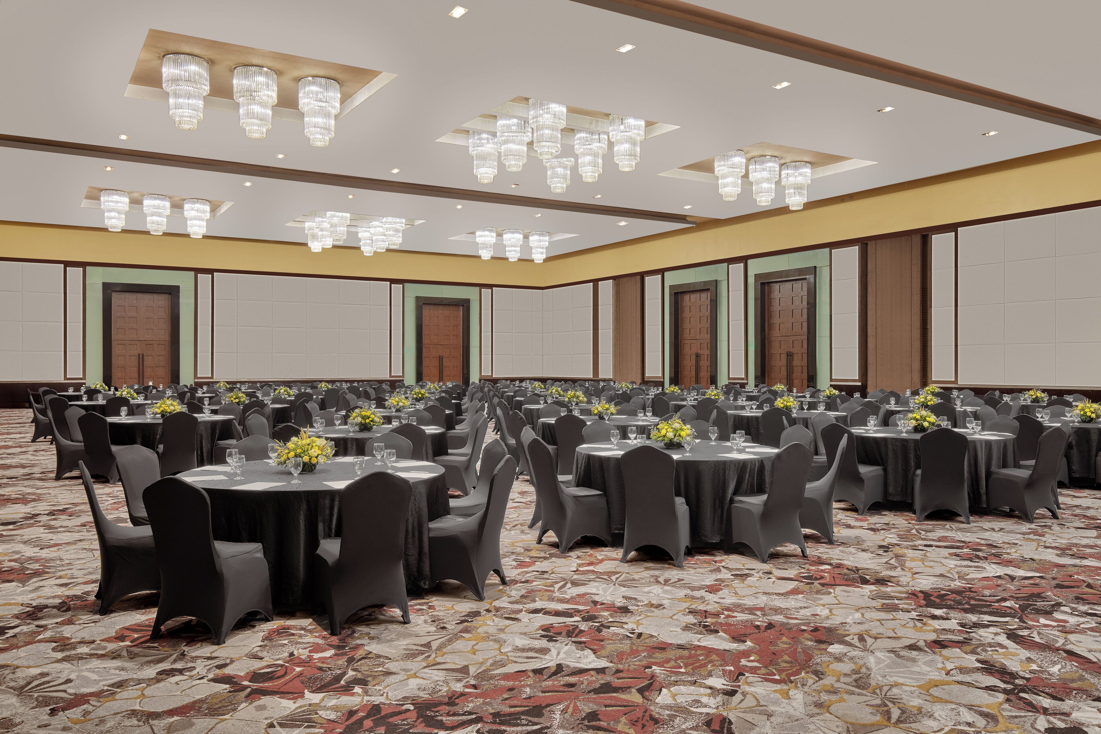 A large banquet hall with round tables, black chairs, and chandeliers.