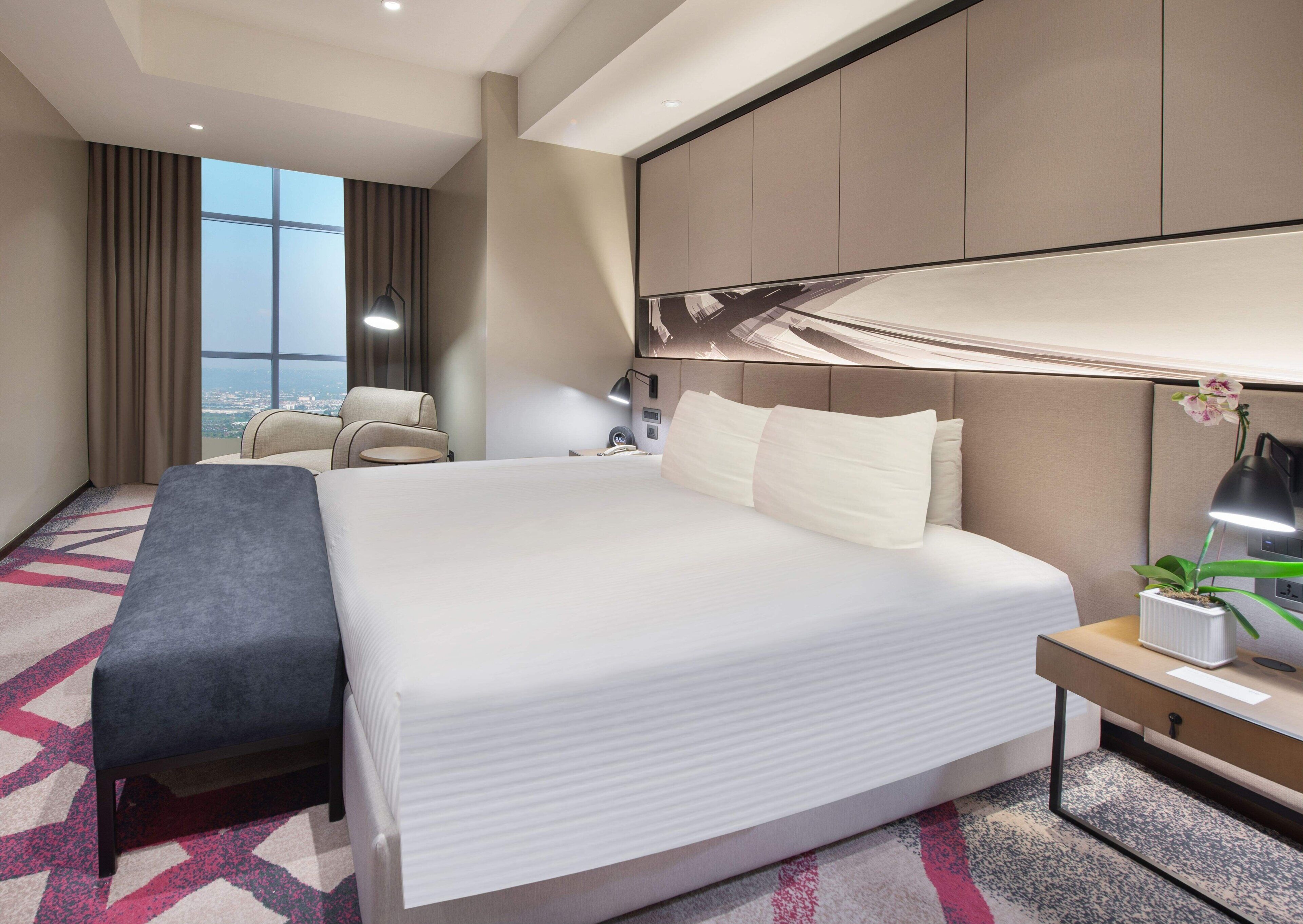 Una habitación de hotel moderna con una cama grande, un escritorio, una silla, una mesita de noche, una lámpara y vistas a la ciudad.