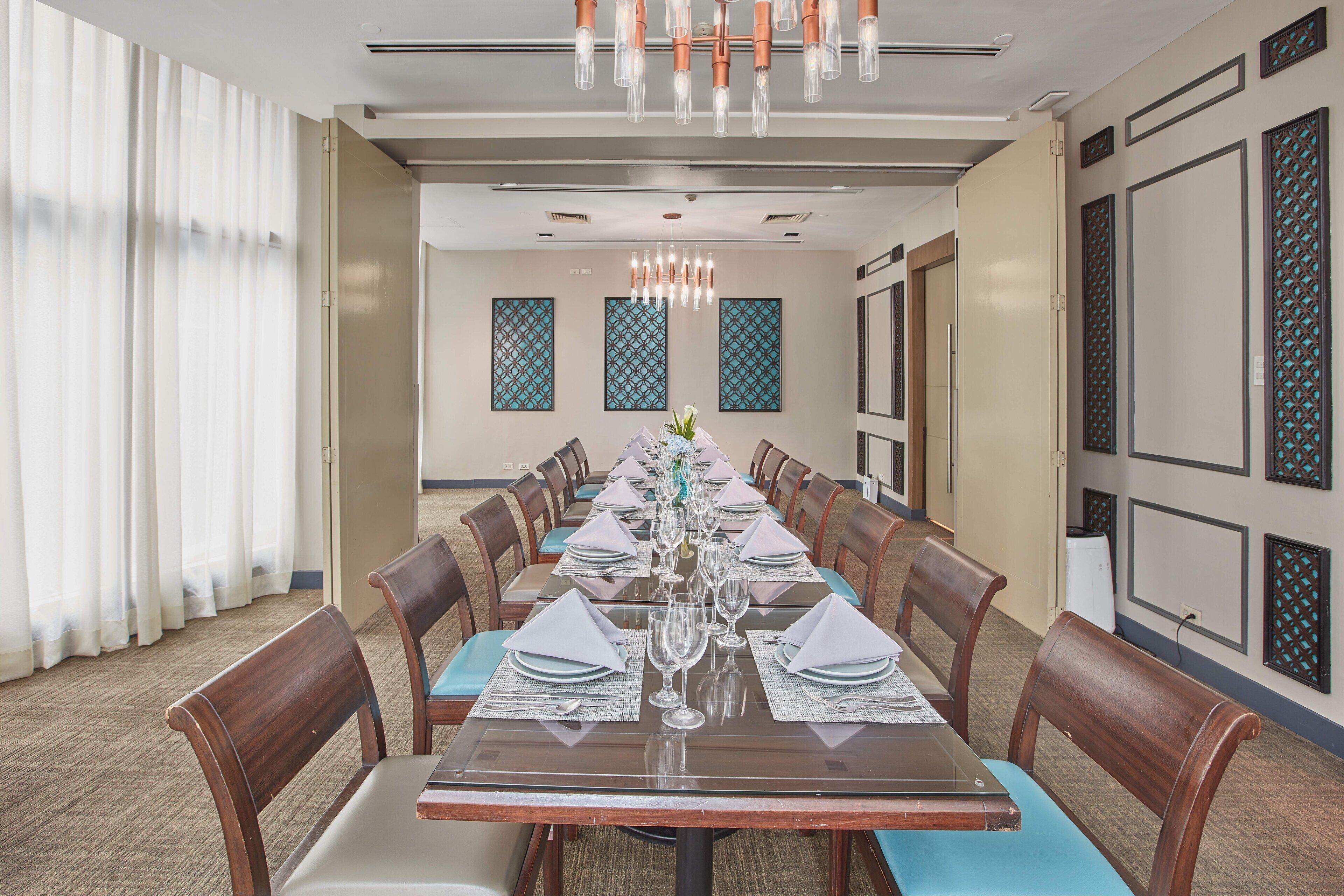 A conference room with a long table set for a meeting, surrounded by chairs, with patterned wall decorations and a chandelier.