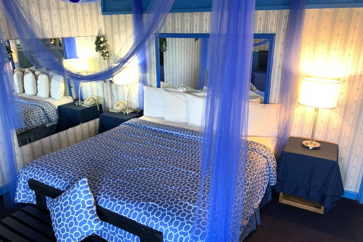 A hotel room with a large bed, a bedside table with a lamp, a mirror, and a window with curtains.