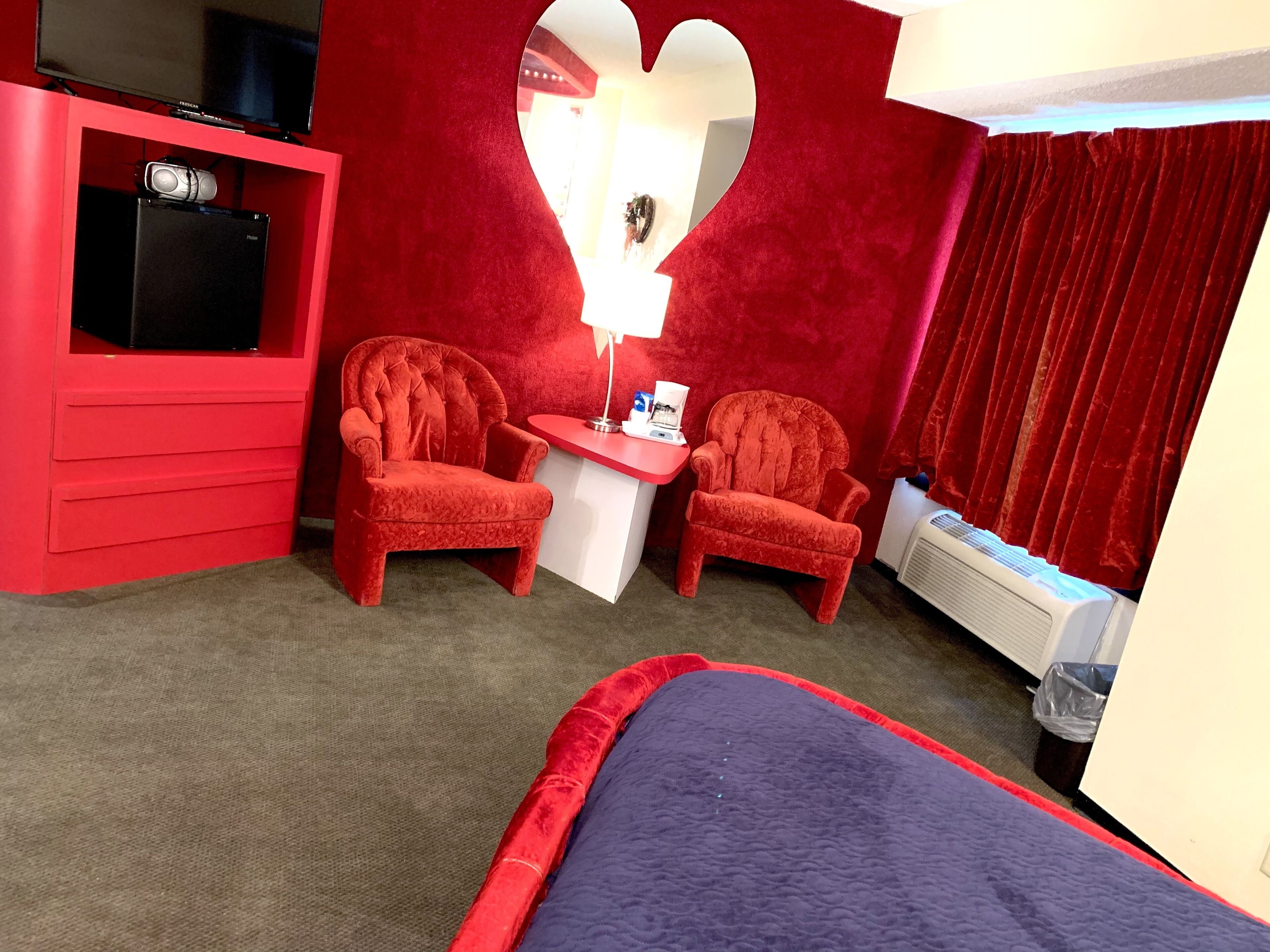 A hotel room with two red armchairs, a small white table, a red wardrobe, a bed with a blue blanket, and a red curtain.