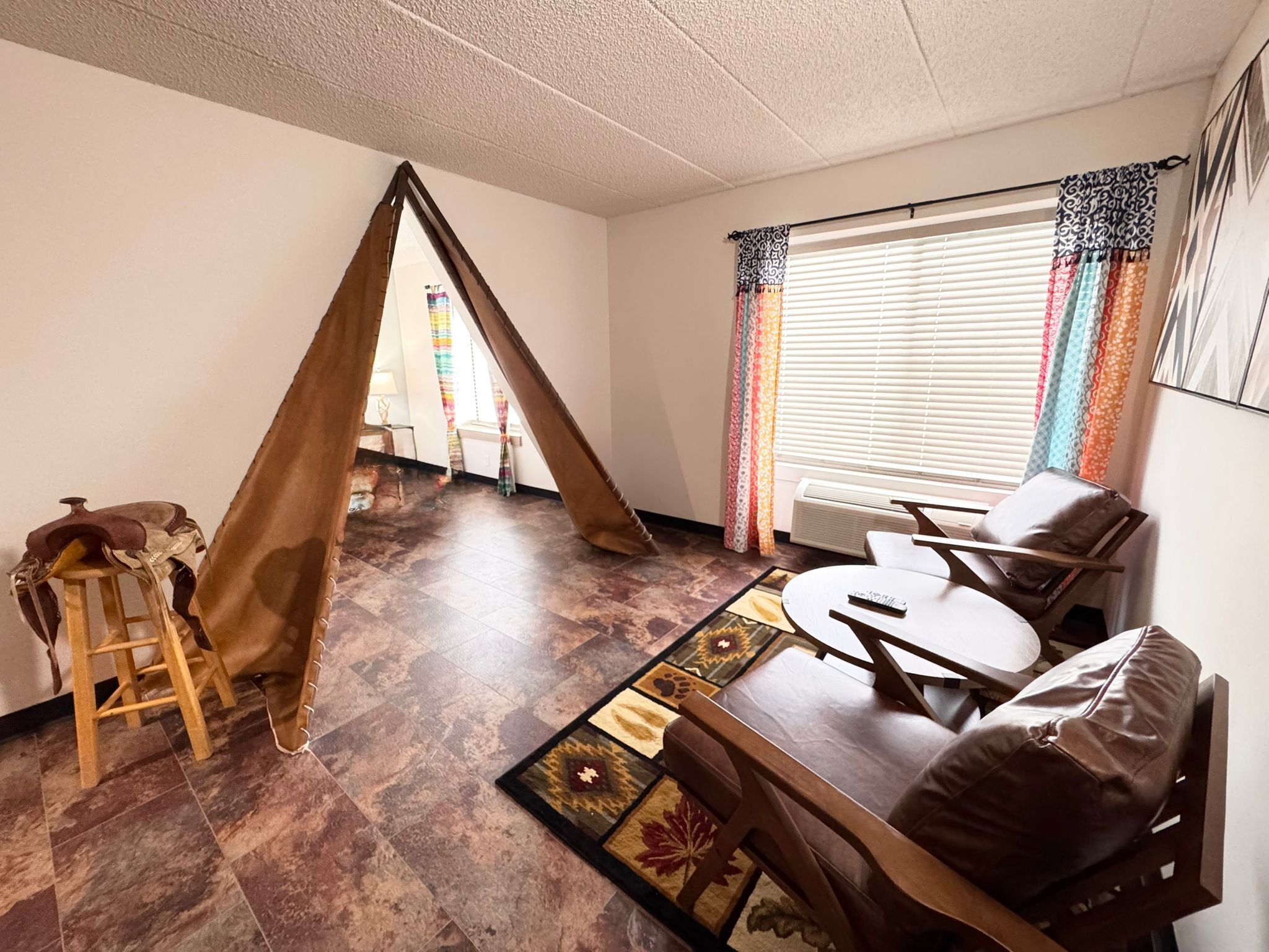 Una sala de estar con un sofá de cuero marrón, una silla de madera, un taburete pequeño de madera y un tapete estampado. Hay una ventana con persianas y cortinas de colores.