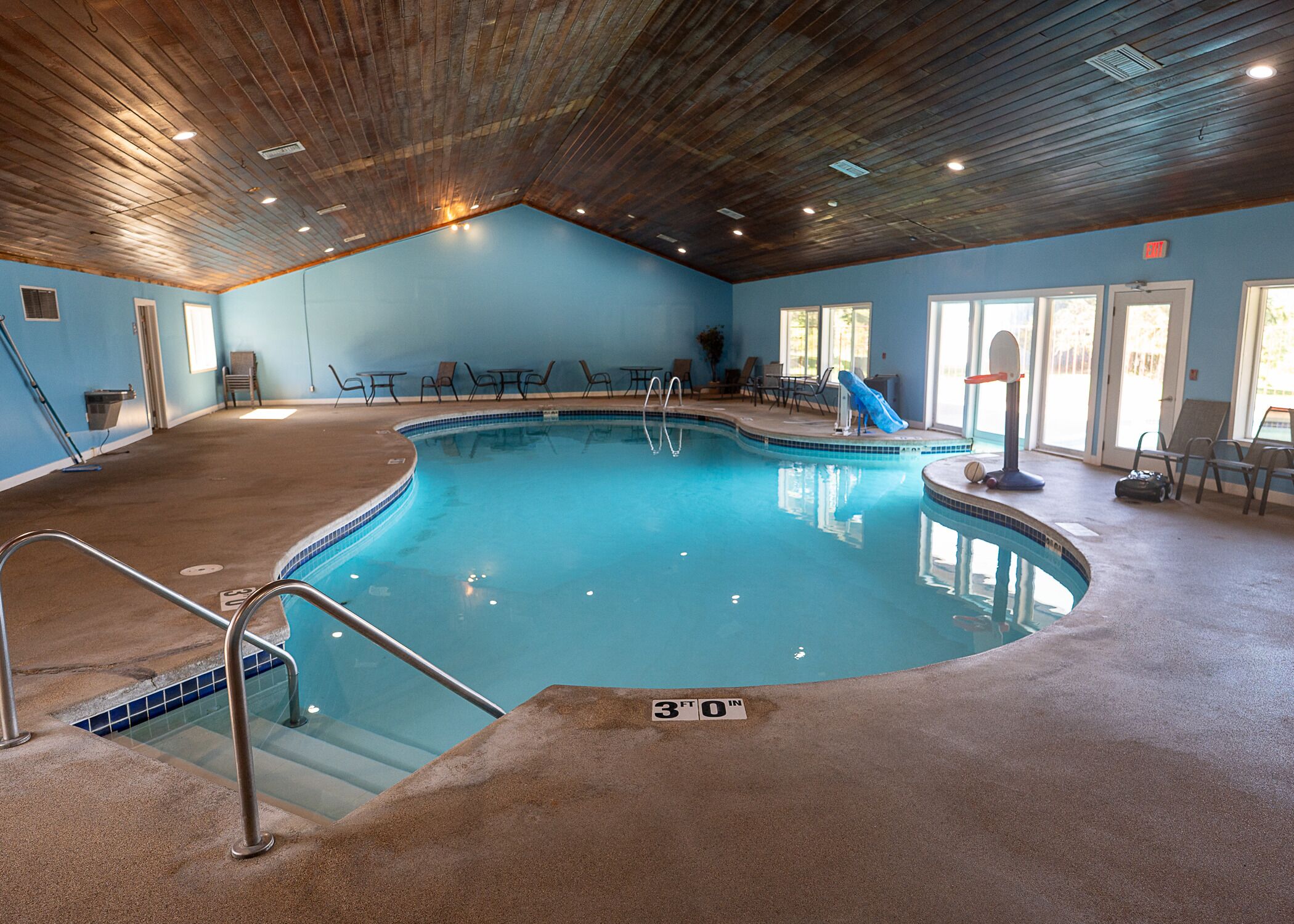 An indoor swimming pool with a 30 ft depth, surrounded by lounge chairs and tables, with a wooden ceiling and blue walls.