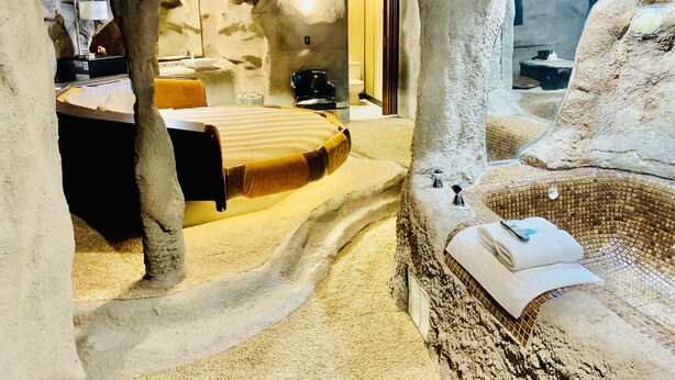 Una habitación con una cama, una mesita y una silla, todo dentro de un espacio que recuerda a una cueva, con paredes texturizadas y suelo de piedra.