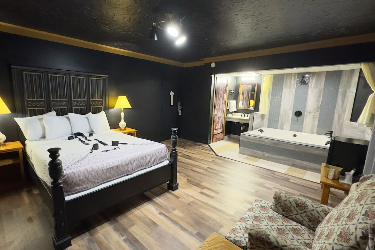 A bedroom with a bed, two bedside lamps, a wooden floor, a chair, and a bathroom with a bathtub and mirror.