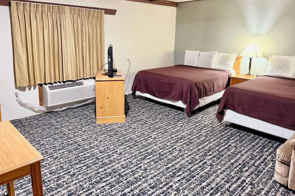 A hotel room with two beds, a wooden nightstand, a desk, a lamp, and a window with curtains.