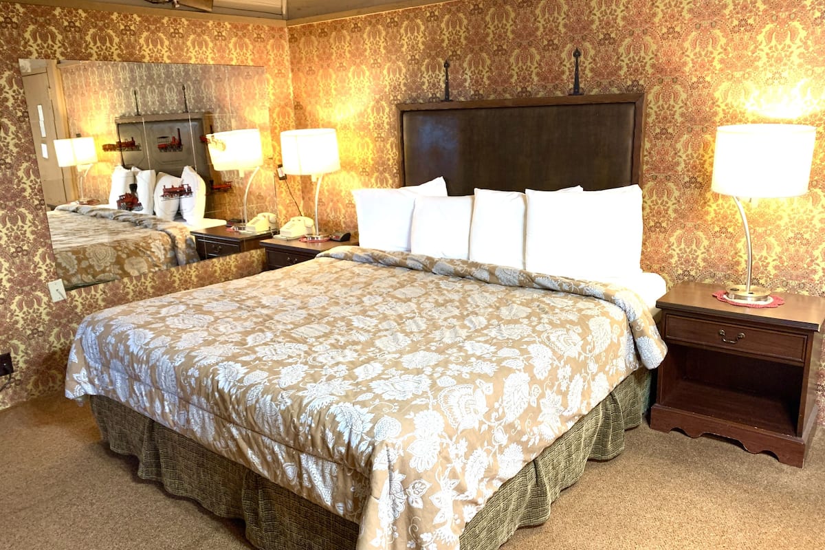 A hotel room with a large bed, two bedside tables, a nightstand, a mirror, and patterned wallpaper.