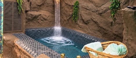 Ett bubbelbad med en vattenfallsfunktion, omgivet av stenliknande väggar och grönska.