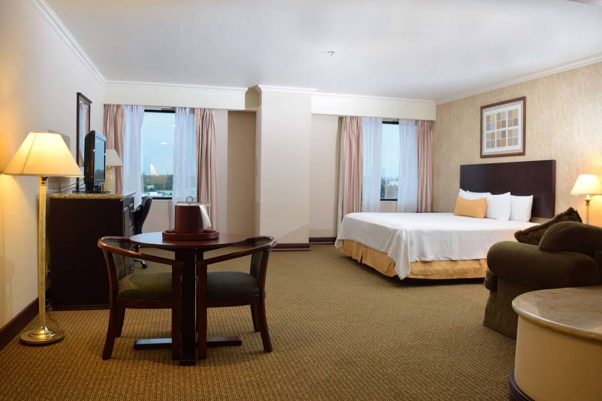 A hotel room with a bed, a desk with a chair, a lamp, and a view of the city through the window.