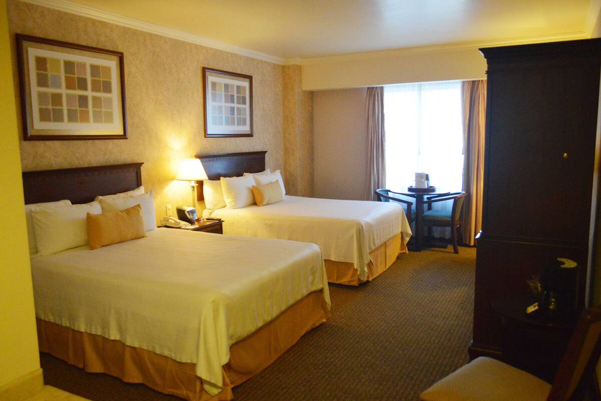 A hotel room with two beds, a desk with a chair, a lamp, and a window with curtains.