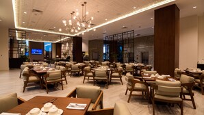 En rummelig hotelrestaurant med flere borde og stole, en moderne lysekrone og et fjernsyn i baggrunden.