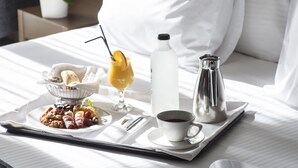 A neatly made bed with white linens, a tray with breakfast items including a coffee cup, a glass of orange juice, and a bottle of water, and a magazine on the bed. - Anatolia (Thessaloniki)