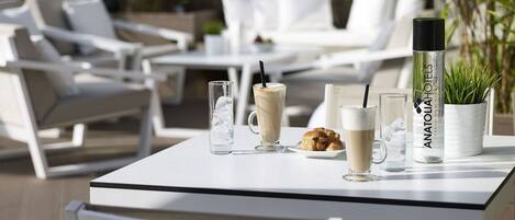Una mesa con bebidas, una botella de Anatolia Hotels y un plato de croissants en un entorno exterior con mobiliario blanco y follaje.