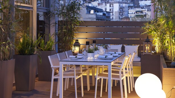 An outdoor dining area with a table set for two, surrounded by potted plants and a view of the cityscape in the background. - Anatolia (Thessaloniki)