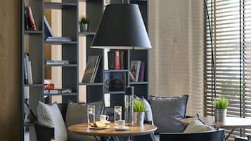 A modern living room with a round wooden table, a black chair, a bookshelf filled with books and decorative items, and a large window with blinds.