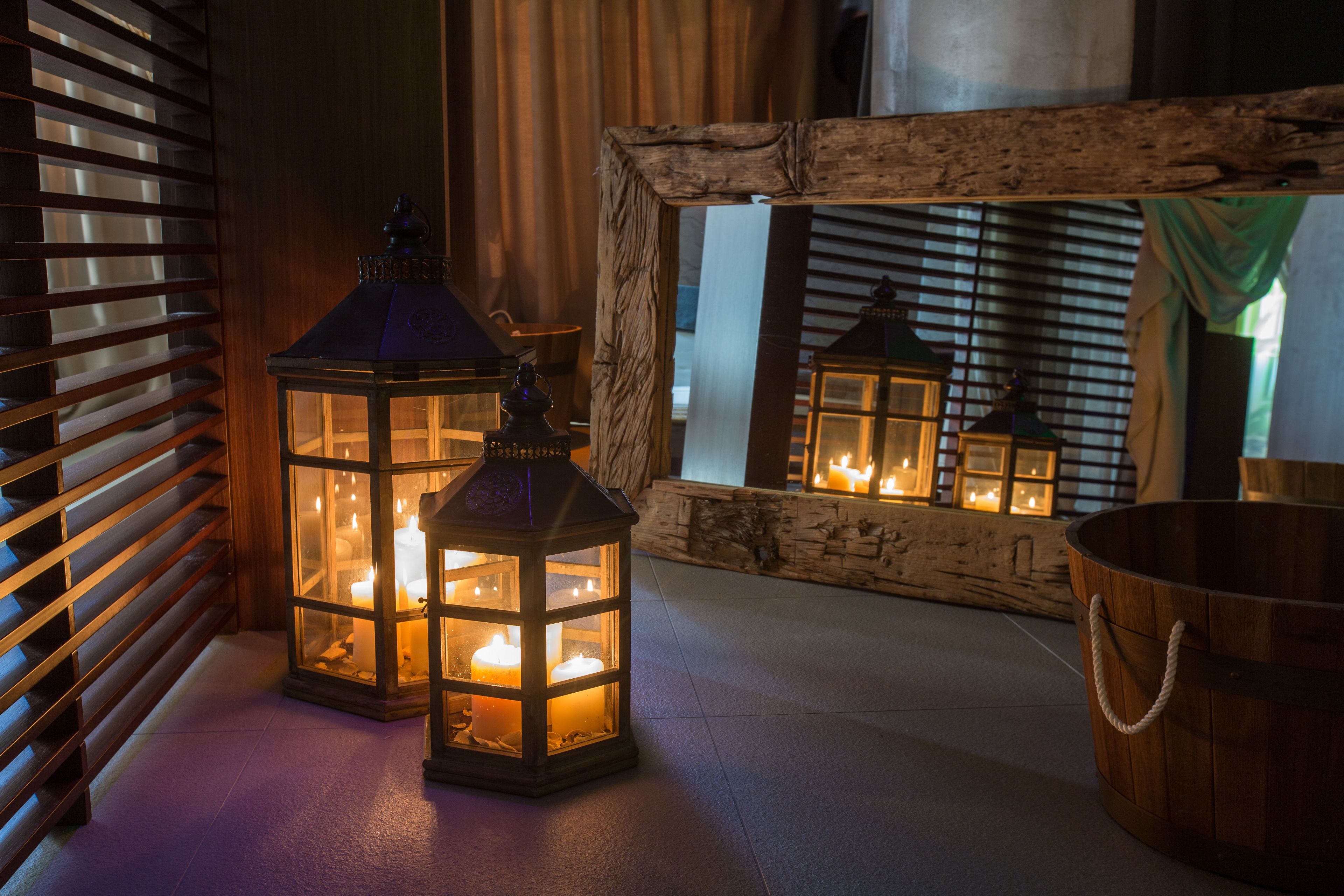 A wooden-framed mirror reflecting two lit lanterns, a wooden bucket, and a wooden bench.